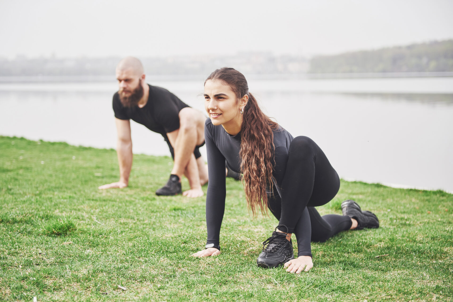 Fitness couple stretching outdoors in park near the water. Young bearded man and woman exercising together in morning